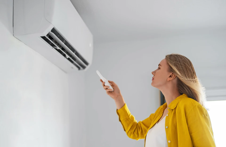 Main tenant une télécommande blanche pointée vers un climatiseur mural dans une pièce moderne avec fenêtres et rideaux clairs
